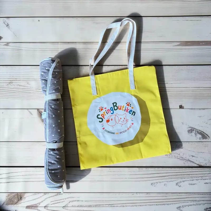 Gul väska med ljusblå handtag och SpringButiken-logotyp, bredvid en hopvikt blå vattentät matta.